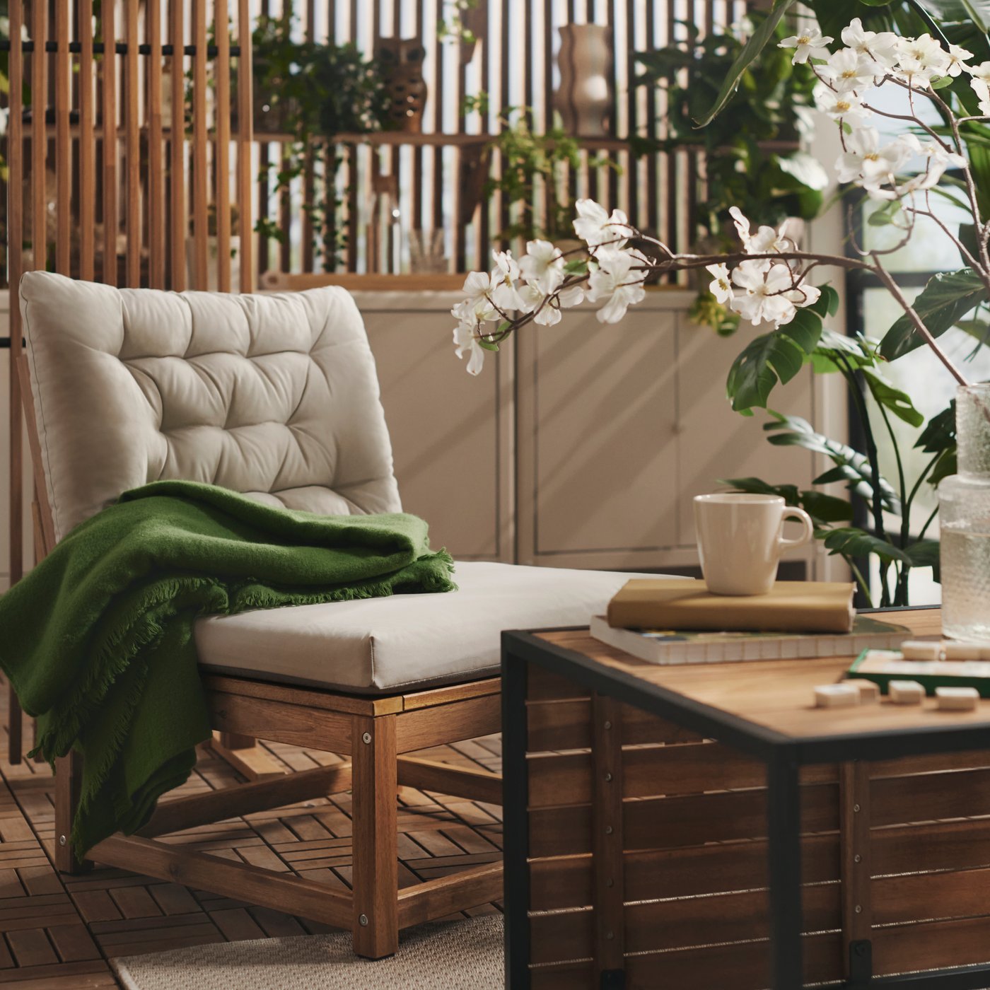 A balcony  with greenery features a light brown stained NÄMMARÖ easy chair with light grey-beige cushions and a wooden table.