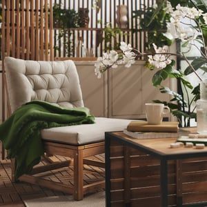 A balcony with greenery features a light brown stained NÄMMARÖ easy chair with light grey-beige cushions and a wooden table.