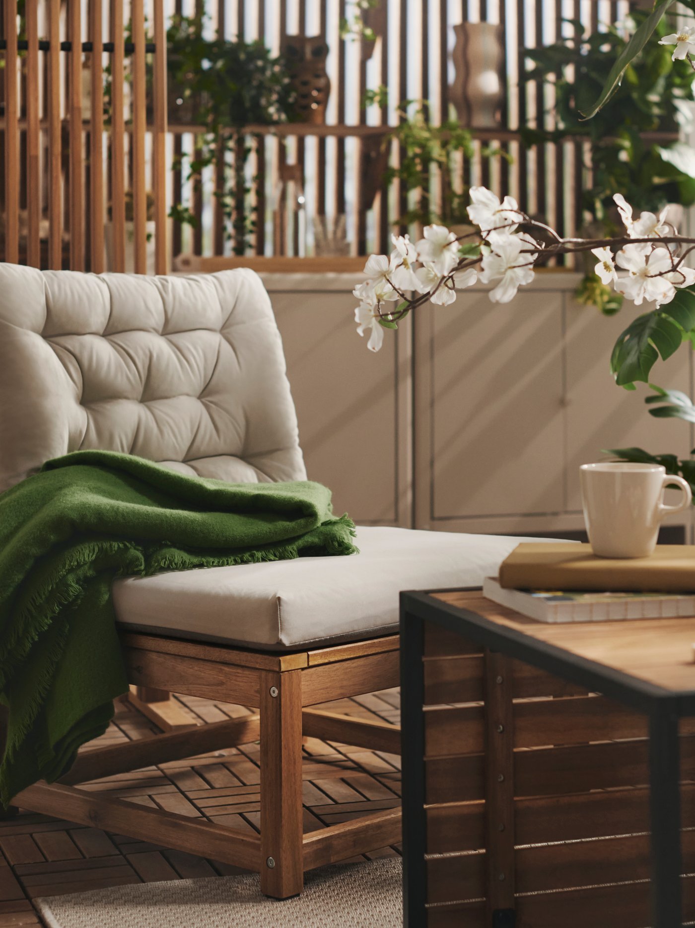 Na balkonie z roślinami znajdują się jasnobrązowy fotel NÄMMARÖ z jasnymi szarobeżowymi poduszkami i drewniany stół.