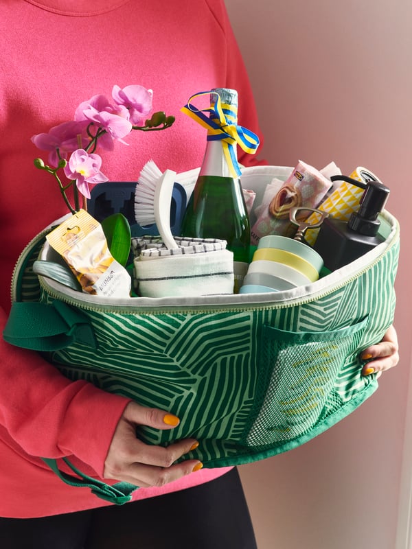 A person holds a green patterned basket filled with beauty products, a small flower arrangement, and assorted toiletries.