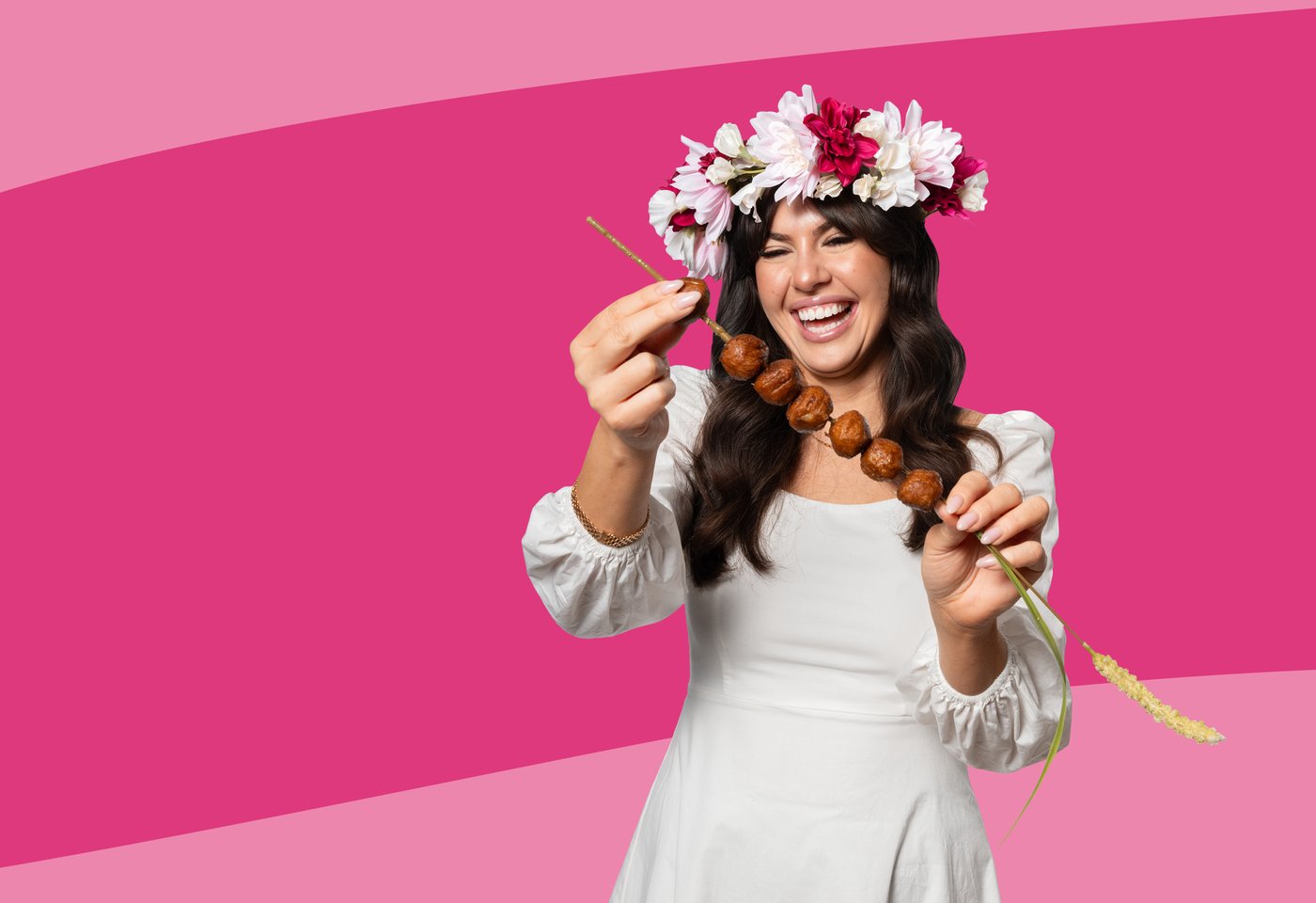 Eine Person mit einem Blumenkranz auf dem Kopf hält vor sich einen Spieß mit Fleisch(los)bällchen.