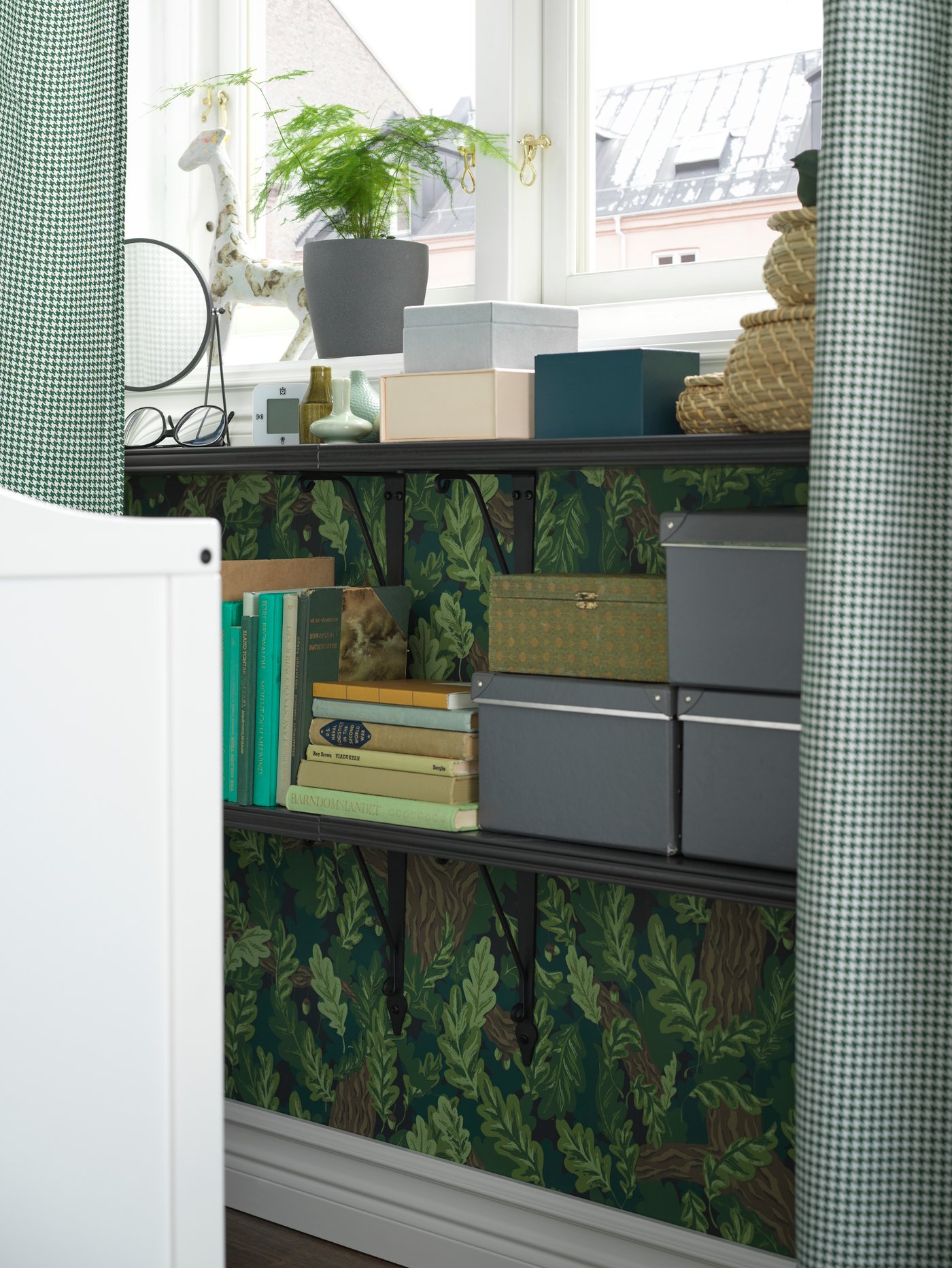 Könyveket, dobozokat és dekorációkat tartó falra szerelhető BERGSHULT/KROKSHULT polcok, egy ORDENSFLY függönyökkel felszerelt ablak alatt.