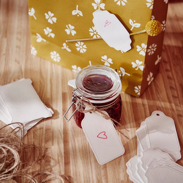A glass jar with a lid filled with jam sits on a worktop. A label with a written greeting is attached to the jar by a string.