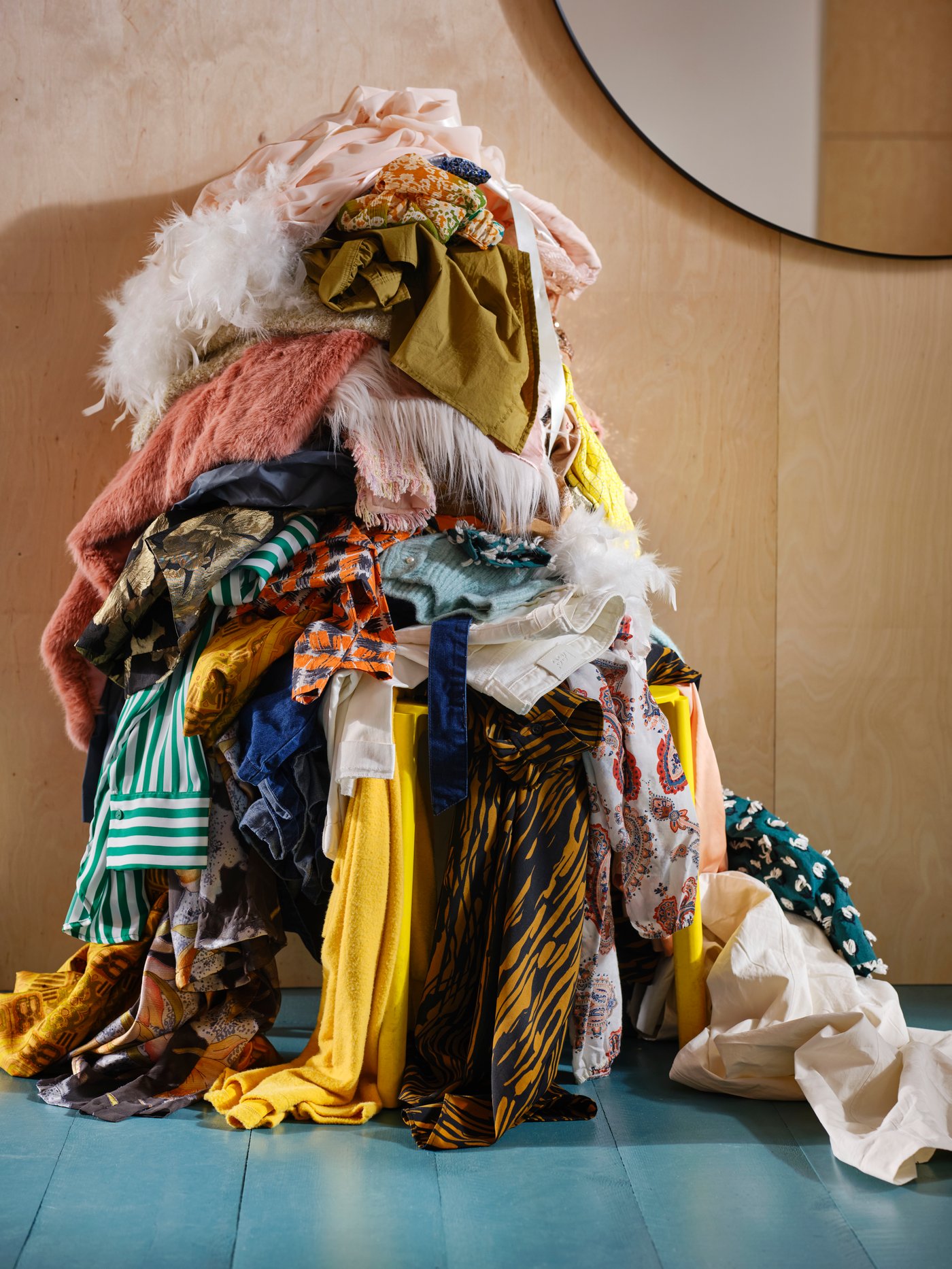 An untidy pile of clothes sits on a yellow chair near a LINDBYN mirror, with many clothes hanging down onto the floor.
