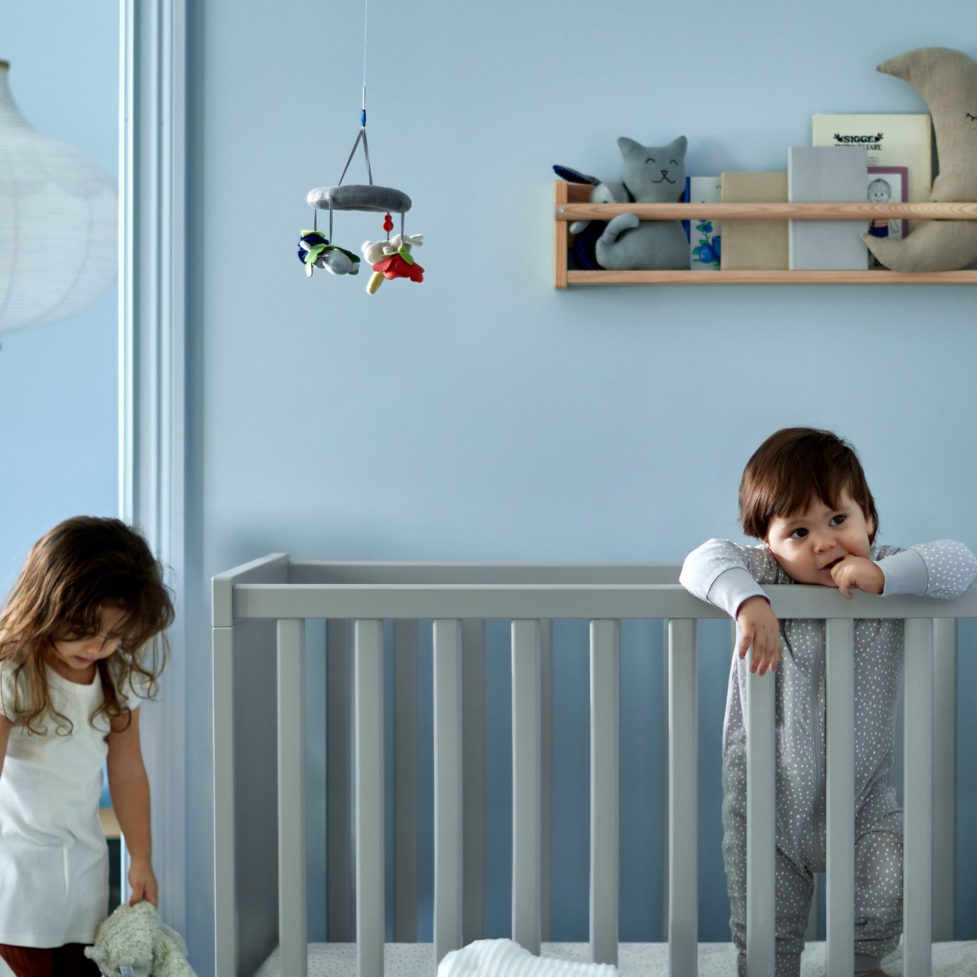 A girl holding two soft toys beside a toddler standing in a grey cot, with a children’s bed with blue bed linen behind.
