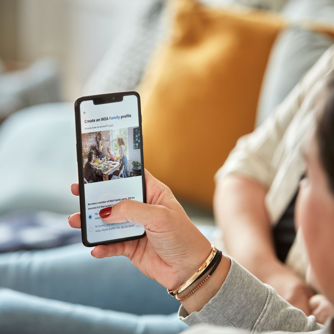 Person håller en smartphone med IKEA Family-registrering på skärmen, sittande i ett ljust vardagsrum med blå fåtölj och senapsgul kudde i bakgrunden.
