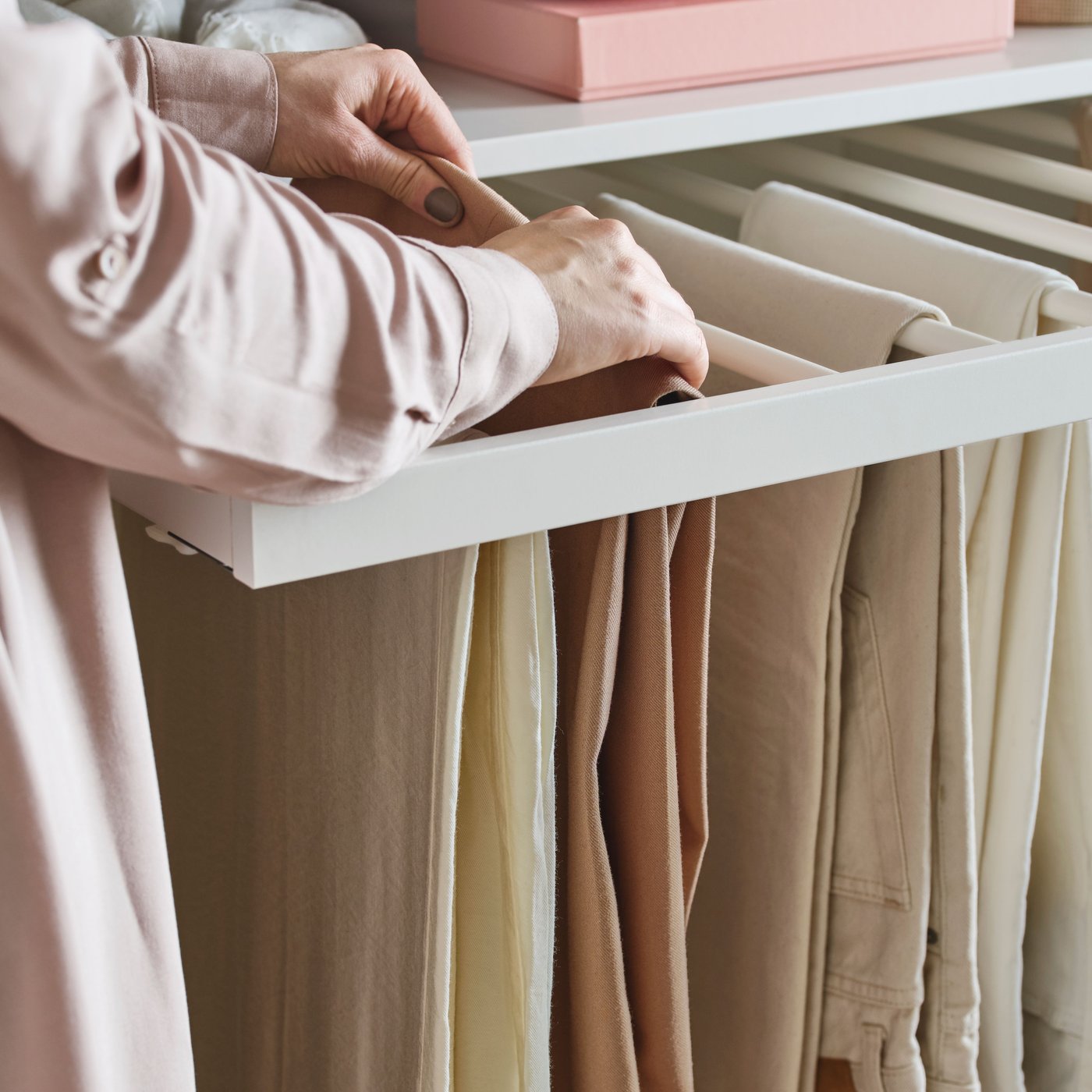 A person in a pink shirt adjusts a pair of beige trousers, one of several on a white KOMPLEMENT pull-out trouser hanger.