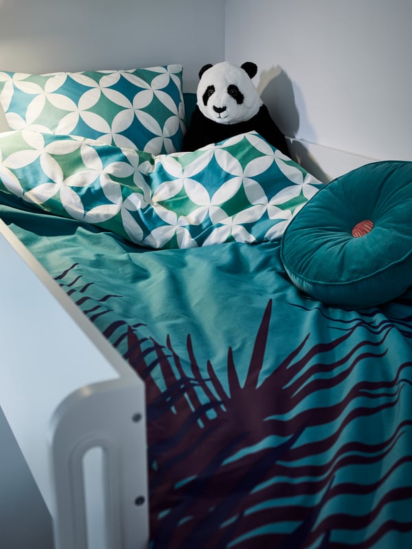 A loft bed with blue and green patterned pillow case and duvet cover, a torquoise round cushion, a panda bear soft toy.