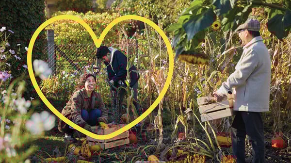 Couple jardinant dans un jardin luxuriant avec un homme portant une cagette en bois. Un logo représentant un coeur apparaît sur l'image en jaune