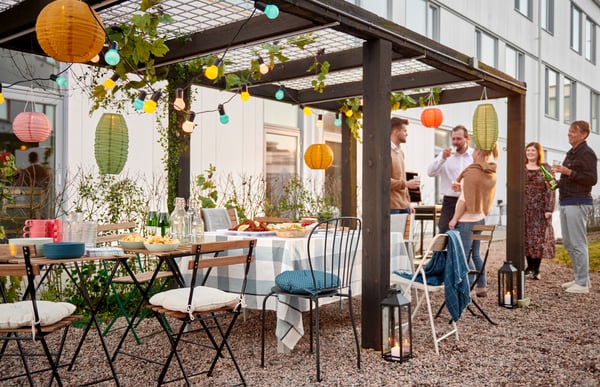 Dans une cour d'immeuble avec pergola en bois décorée de lumignon et table extérieure dressée comme pour une fête, des personnes ont l'air de participer à une fête et discutent debout.