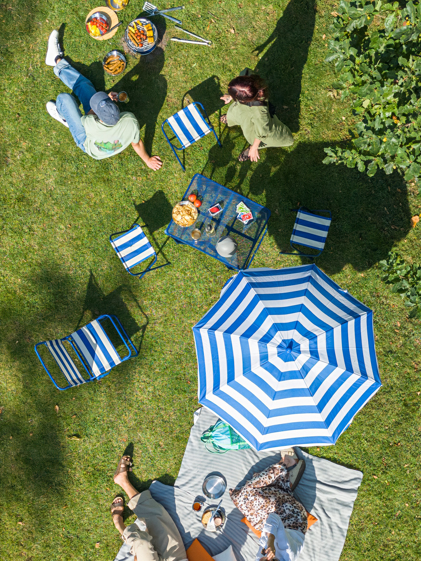 Friends have a barbecue in the park, relaxing on the grass with a STRANDÖN table, chairs, stools, and parasol.
