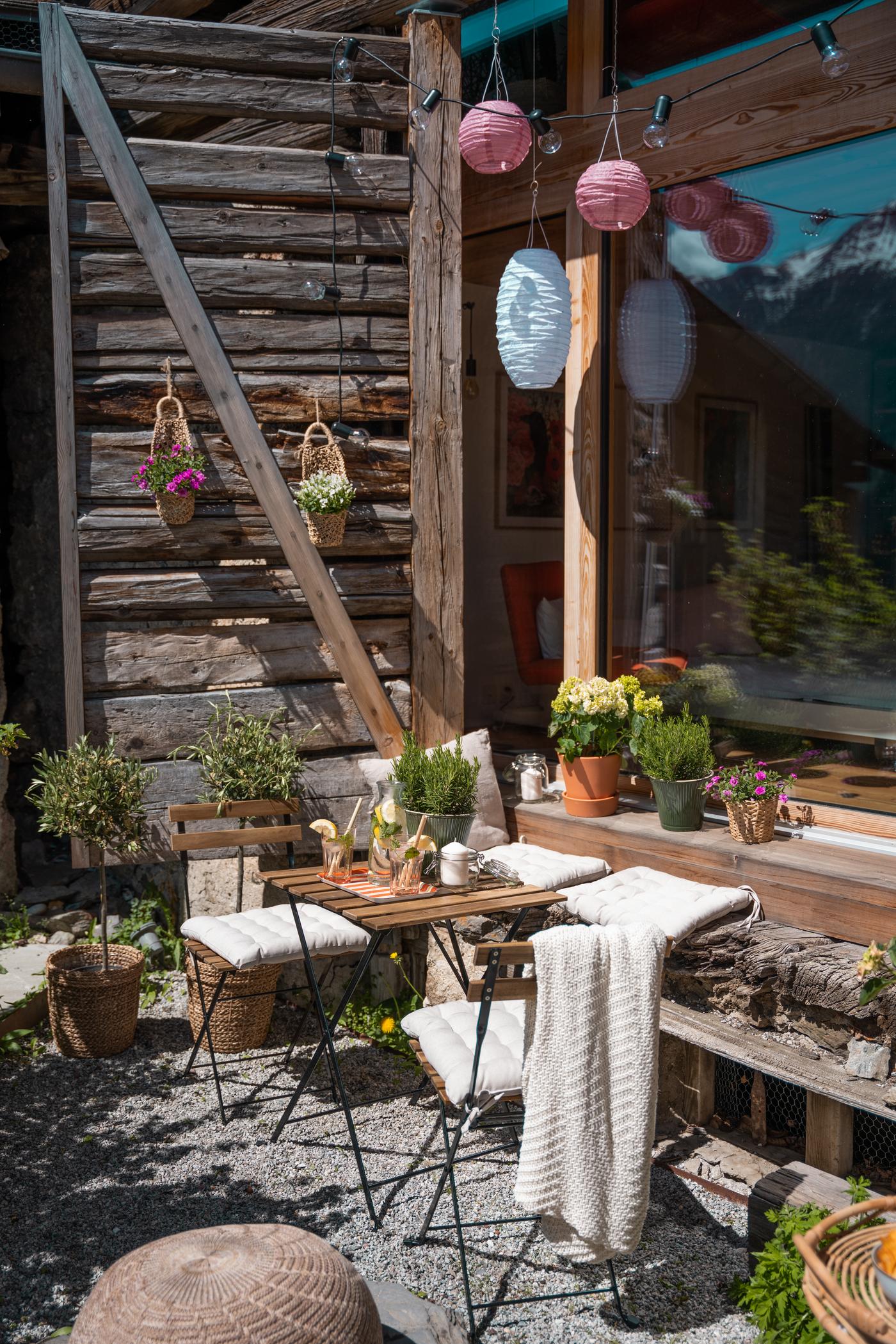 Gemütliche Gartenterrasse mit Holztisch, Pflanzen und Laternen vor einem rustikalen Chalet in den Alpen.
