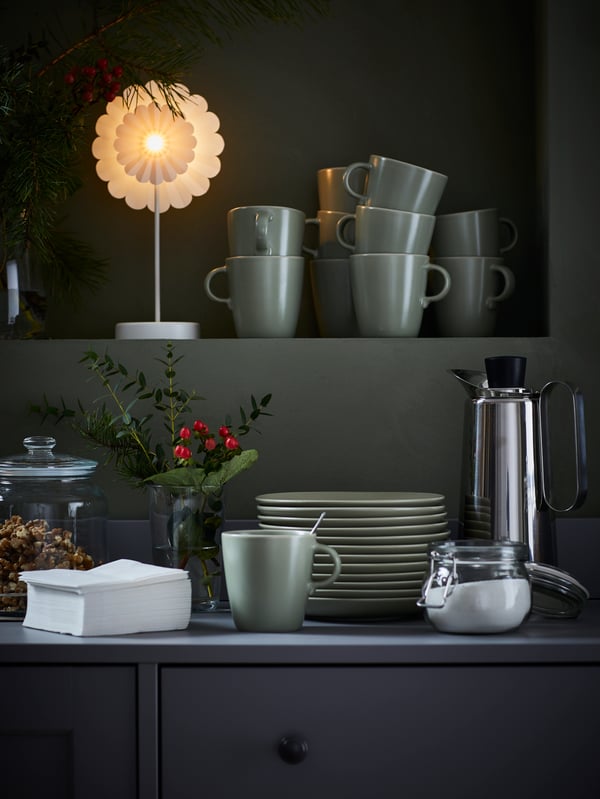 Stacks of matt-green FÄRGKLAR side plates and mugs framed by coffee-drinking accessories and decorations.