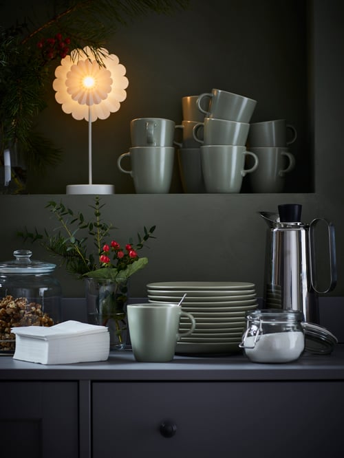 Stacks of matt-green FÄRGKLAR side plates and mugs framed by coffee-drinking accessories and decorations.