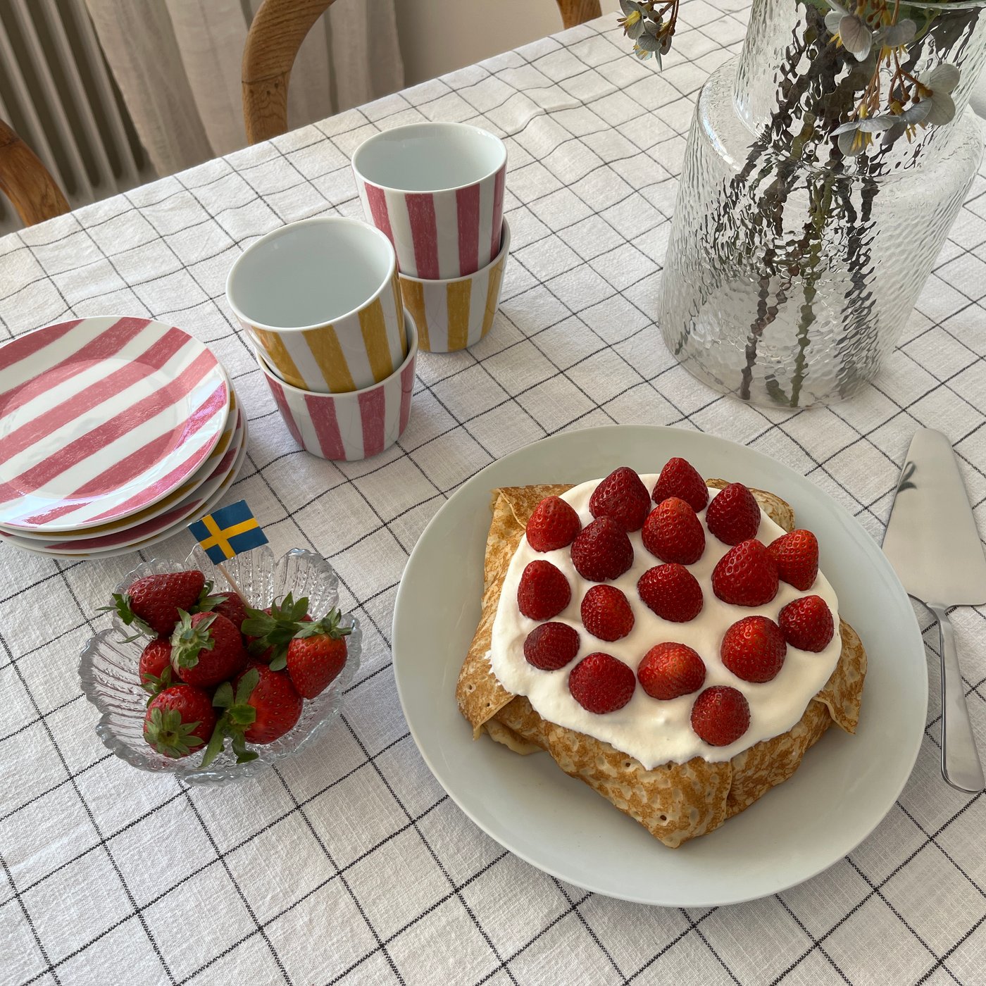 På ett bord med rutig duk syns en tallrik med en pannkakstårta, färgglatt randigt porslin, en skål med jordgubbar och en vas med blommor i.