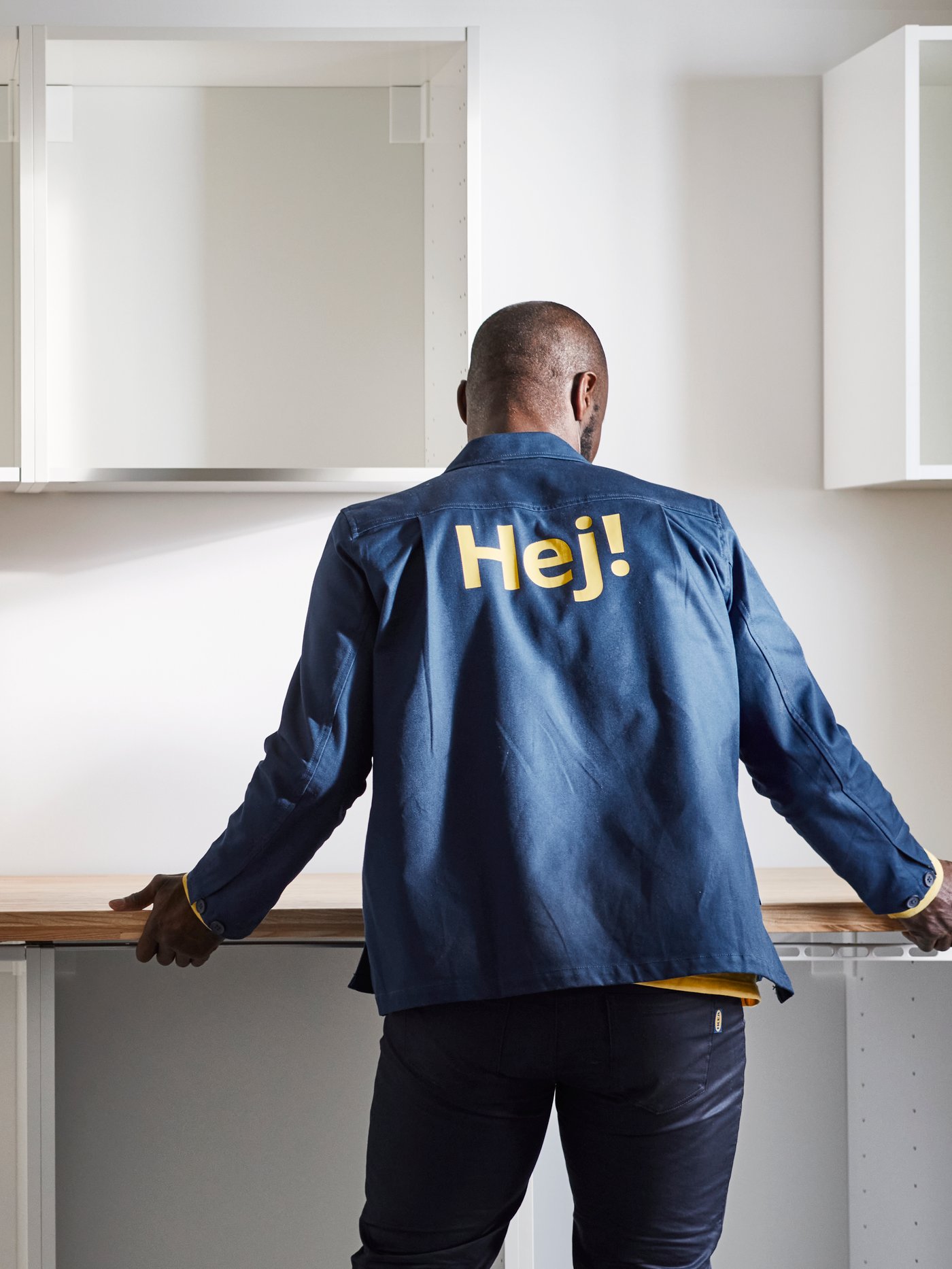 A person stands in a bright kitchen in front of a countertop, wearing a blue jacket with the word “Hej!” printed on the back.