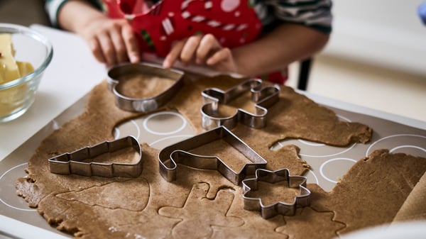 Närbild av en person som använder pepparkaksformar för att stansa figurer i en utkavlad deg på ett bakbord. Formarna har olika former, bland annat hjärta, gran, blomma och pepparkaksgubbe. Personen bär ett rött förkläde med vita detaljer.