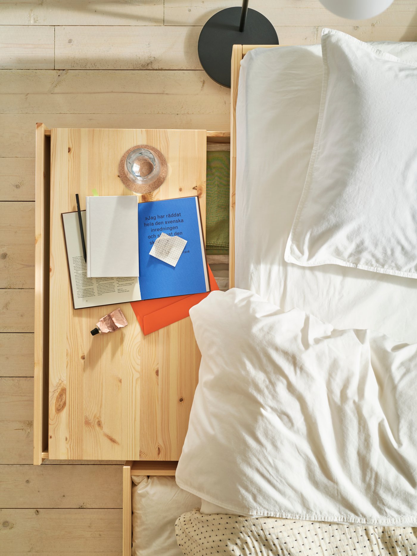 Blick von oben auf ein Bett mit weißen Laken, eine herausgezogene Schublade, ein Tablett aus Naturholz, ein Buch und ein Glas.