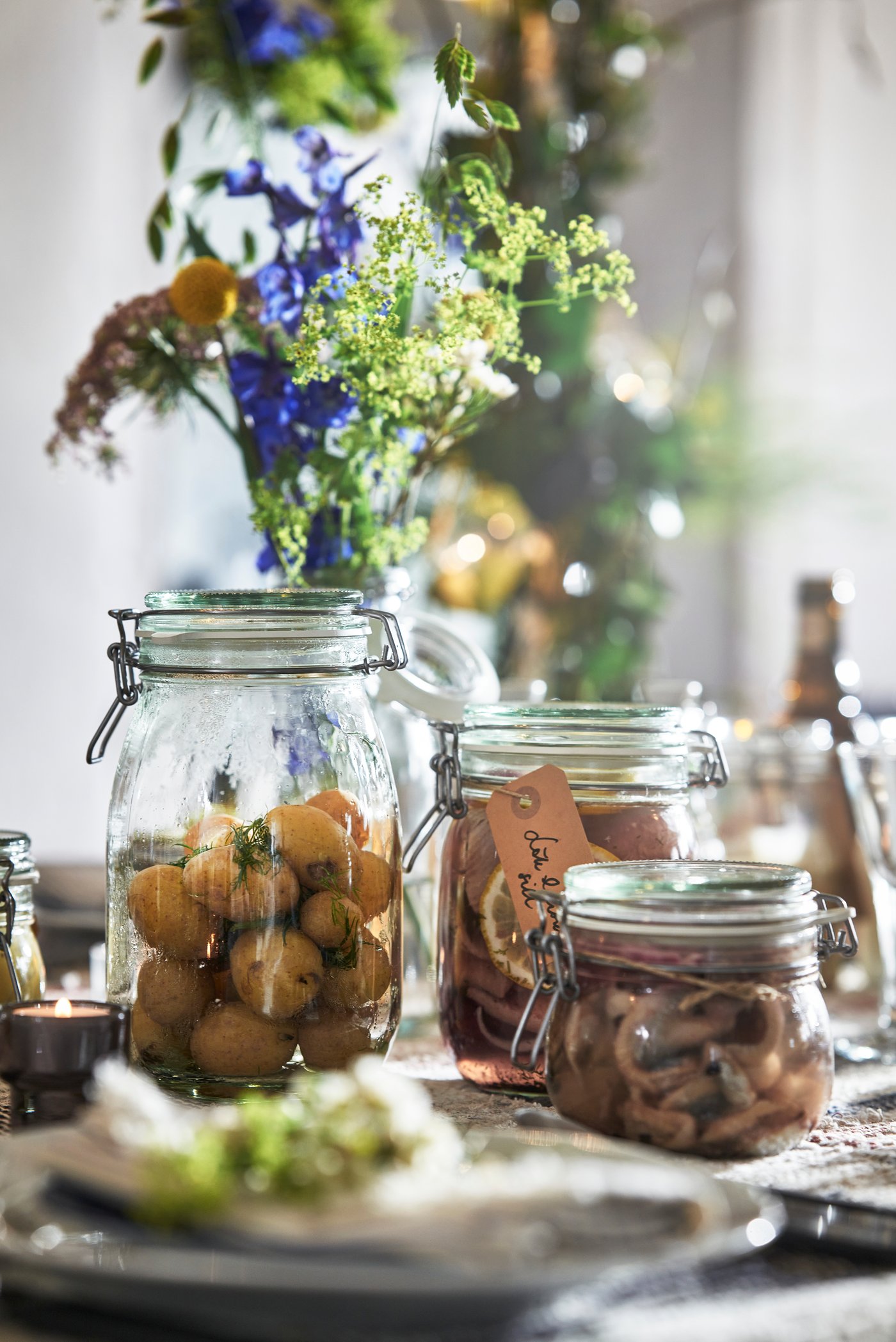 Närbild av ett dukat bord med glasburkar fyllda med små bakverk och nötter; en burk är dekorerad med blå och gröna blommor, och runtom syns grön dekoration, textilier och mjukt naturligt ljus i bakgrunden.
