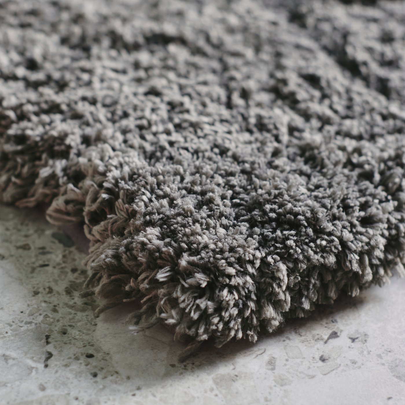 Pohľad zblízka na roh tmavosivej kúpeľňovej predložky ALMTJÄRN a jej bohatý, mäkký vlas, ktorá leží na sivej podlahe s mramorovým vzorom.
