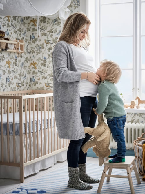 A child standing on a VILTO stool in front of their pregnant mum who is touching their face. A SNIGLAR cot is behind them.