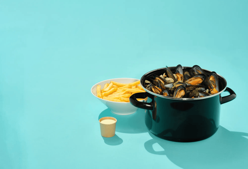 Mosselen in zwarte pot met frietjes en saus op blauwe achtergrond, eenvoudig gepresenteerd.
