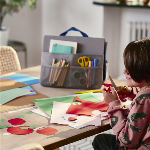 Tavolo con fogli colorati, cartoncini, pennarelli, forbici e altri materiali creativi; bambina intenta a ritagliare delle forme di carta rossa e verde.