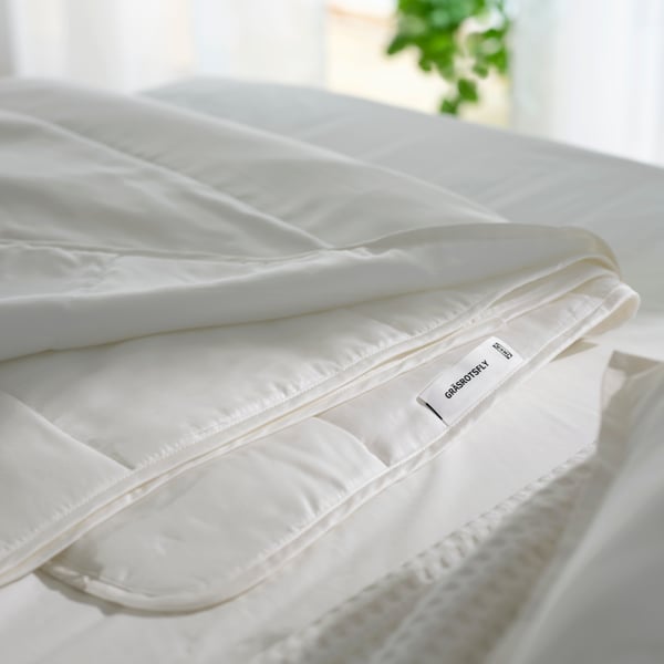 Een netjes opgevouwen GRÄSROTSFLY-dekbed ligt op een bed naast een raam met een groene plant.