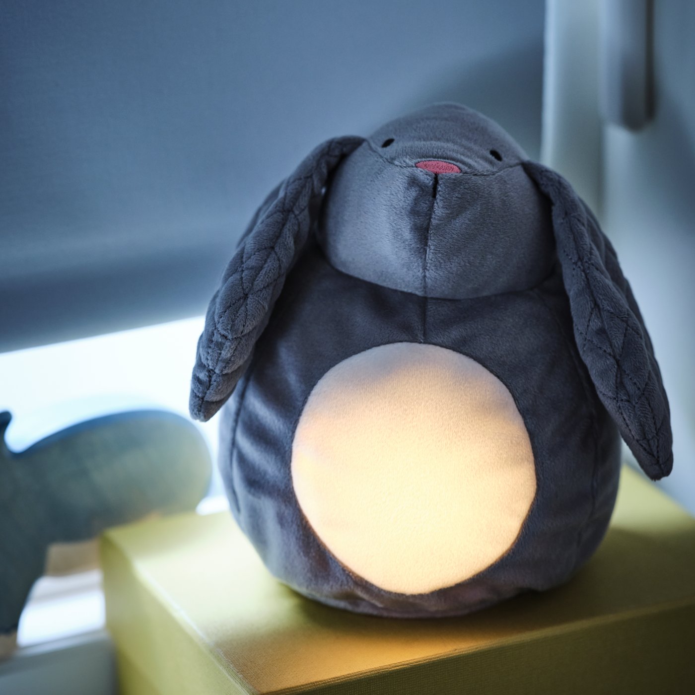 A grey PEKHULT soft-toy rabbit with an LED night light sits next to a pulled-down roller blind, emitting a soft glow.