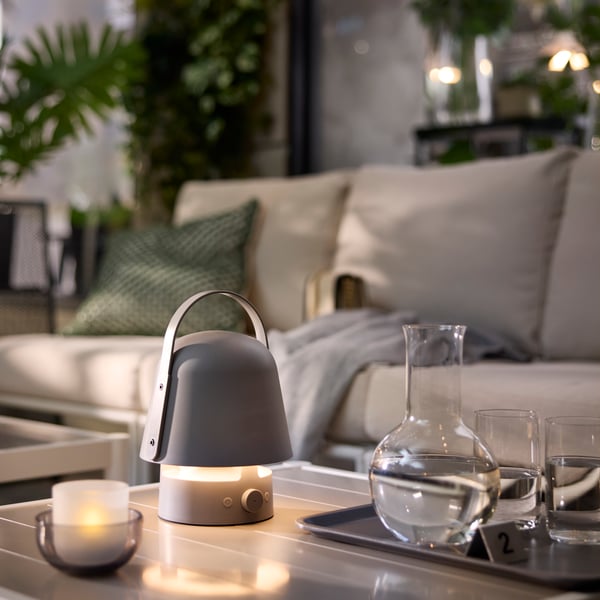 A grey VAPPEBY bluetooth speaker lamp sits on a table, surrounded by plants, glasses, and candles in an outdoor café setting.