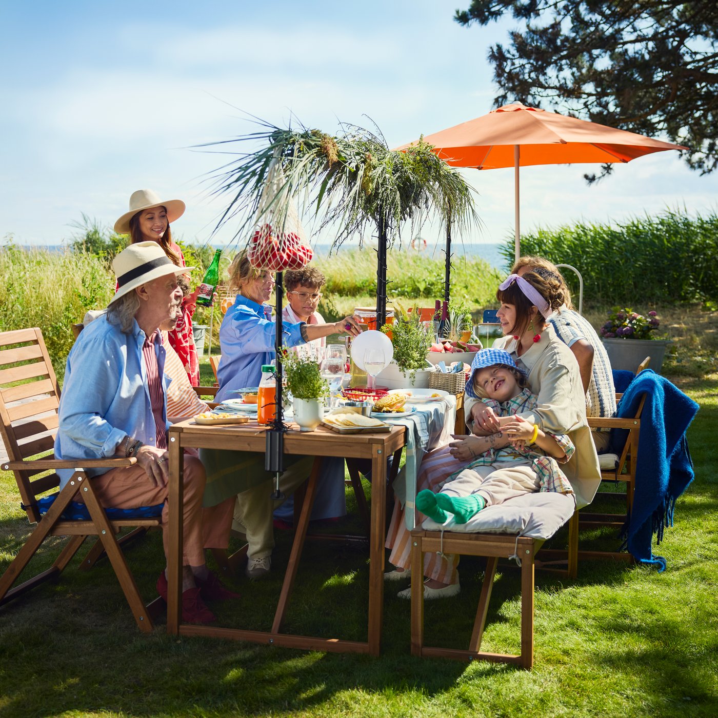 A family is sitting at a wooden garden table in a yard with a wide view, enjoying good food.