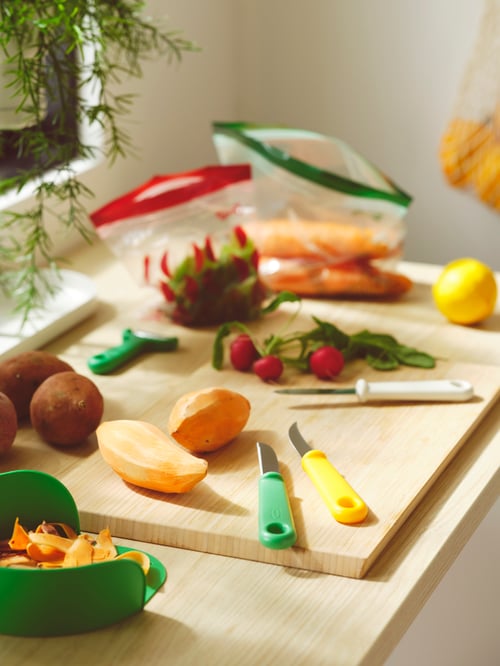 Un comptoir d’armoire sur lequel se trouvent trois couteaux d’office, un économe, des légumes dans des sacs refermables et d’autres légumes sur une planche.