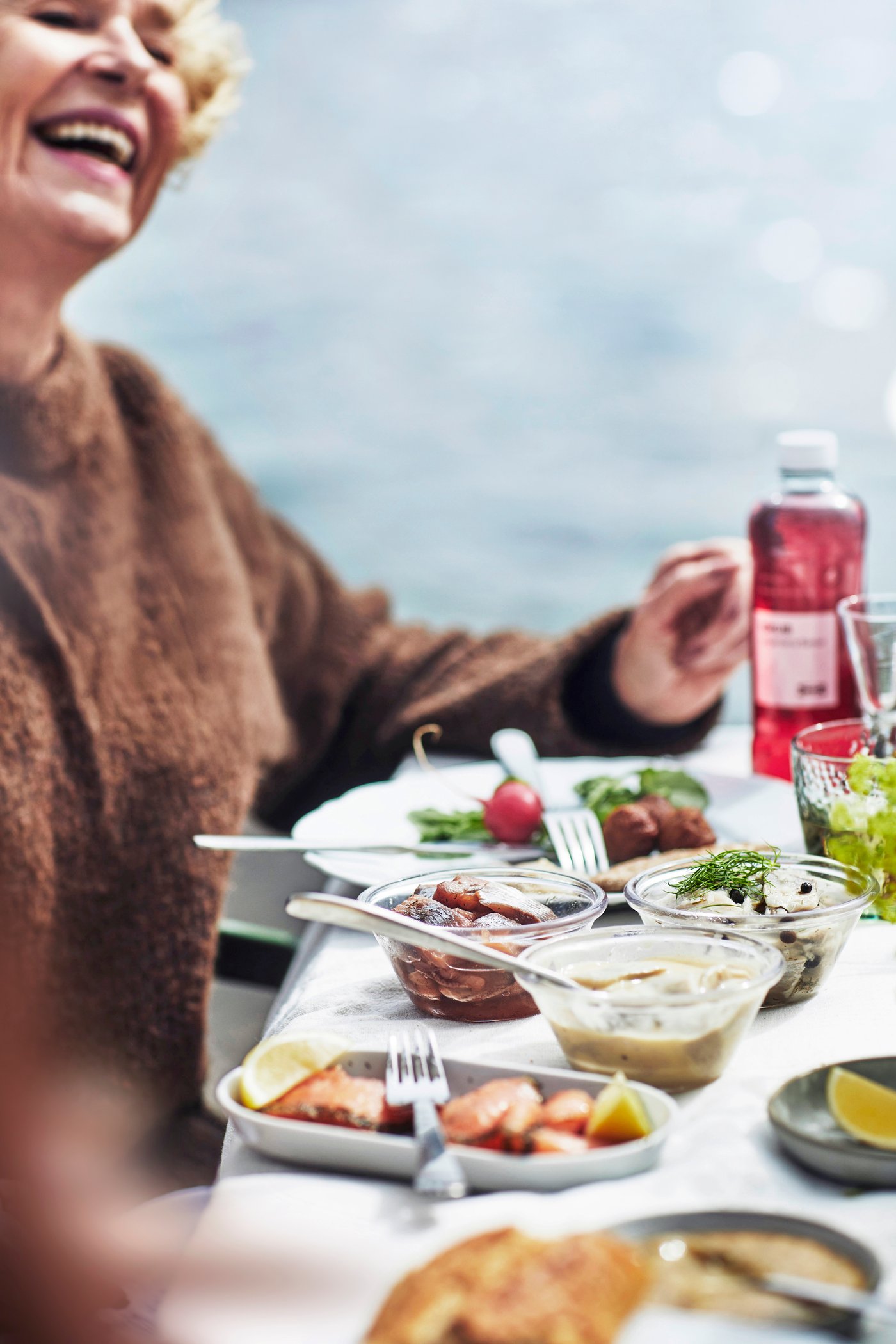 A laughing woman sits at a set table with food and bowls of various types of herring in front of her.