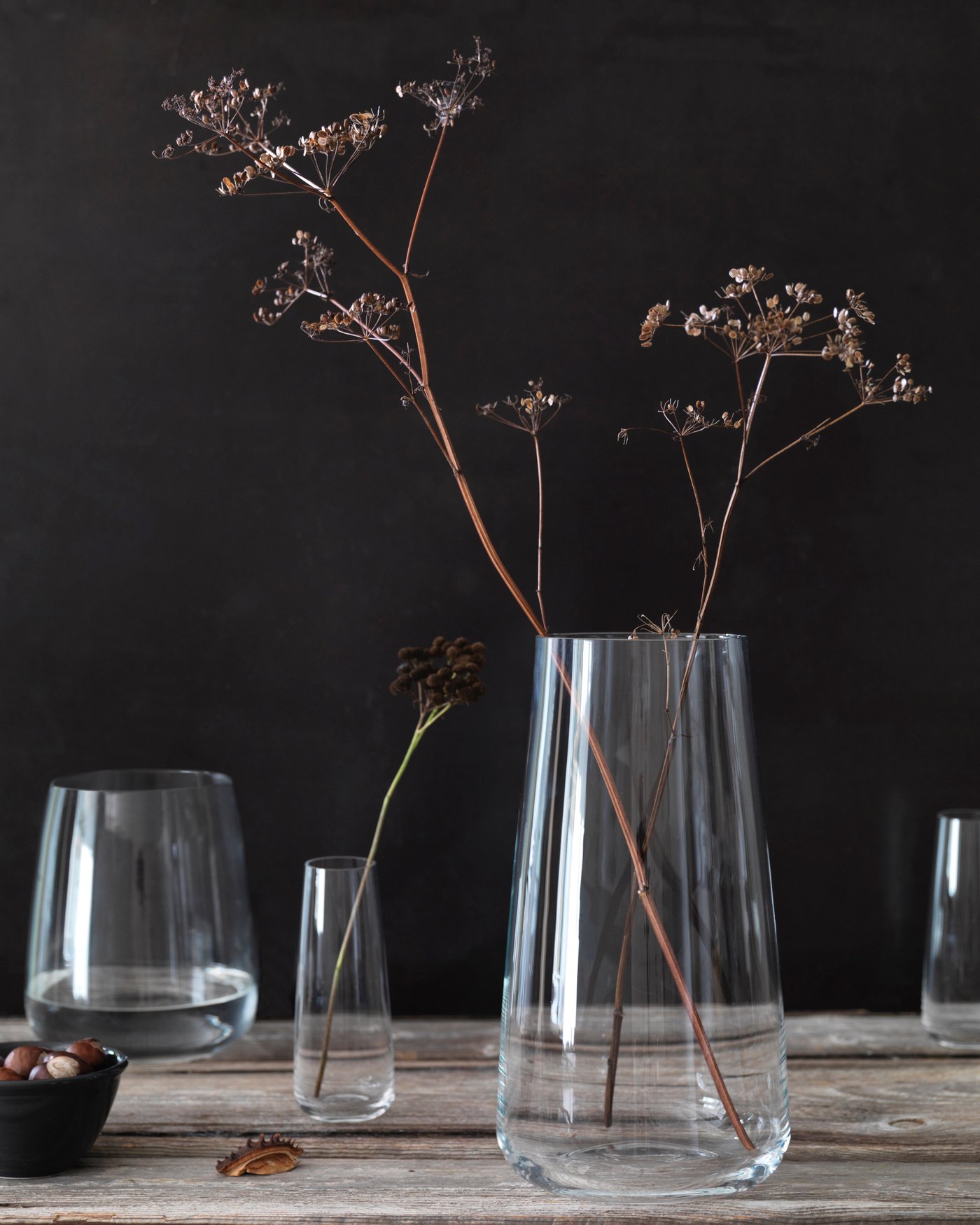 En minimalistisk stilleben med tre glasvaser i olika storlekar med torkade grenar, samt en liten svart skål med ekollon på en träyta mot mörk bakgrund.