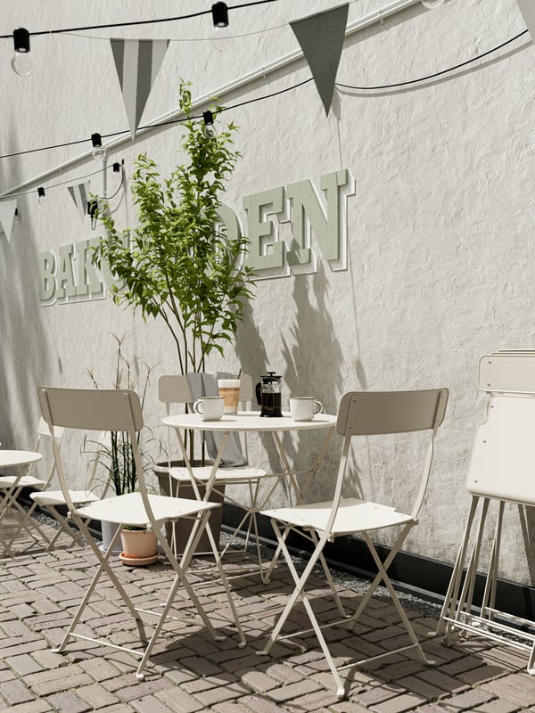 A beige outdoor dining set with coffee on the table and three chairs. Folded chairs lean against a white wall.