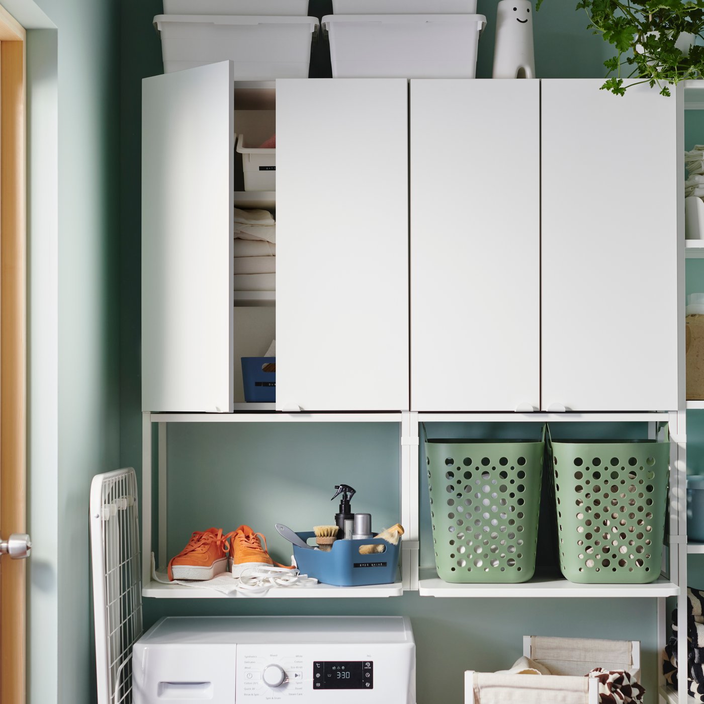 Une boîte UPPDATERA ouverte, gris-bleu, contient des outils pour nettoyer les chaussures, à côté d'une paire de chaussures, sur une étagère murale profonde au-dessus d'une machine à laver.