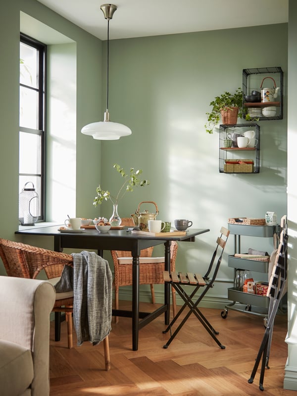 A dining nook in a corner, wit a drop-leaf table with tea on it, rattan armchairs, and a white pendant lamp.
