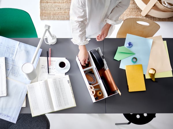 A pair of hands picking an item from a white desk organiser on two joined table tops with work papers and a white work lamp.