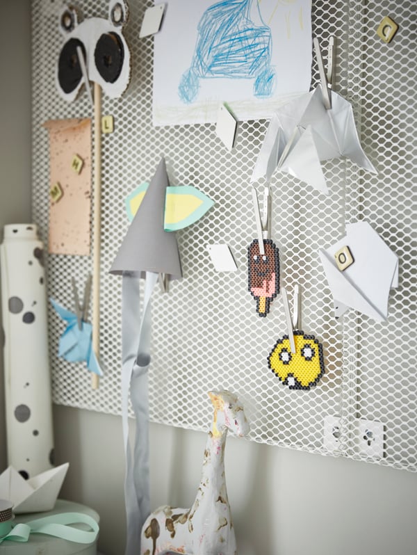 A white, wall-mounted SÖDERGARN memo board with magnets and clips, and attached on them are children’s crafts and drawings.