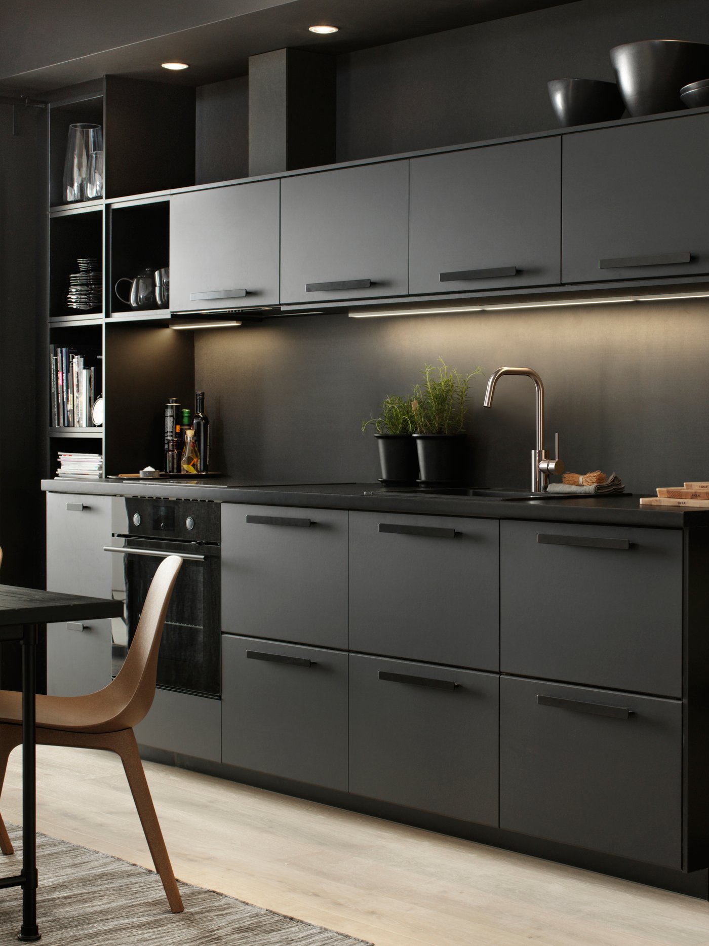 A sleek black kitchen with lighting under the top cupboards. Two black pots with plants inside stand on the counter.