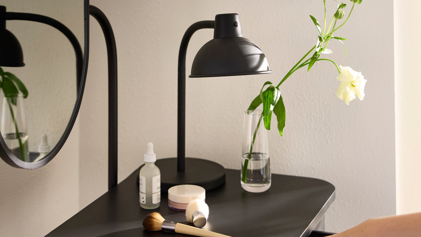 Close-up of GRÅFJÄLLET dressing table with books, make-up and SKURUP work lamp. A hand opening drawer to reach something.