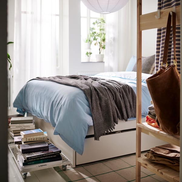 VIHALS bed frame with two storage boxes in a bright bedroom with stacked books, soft bedding and light from tall windows.