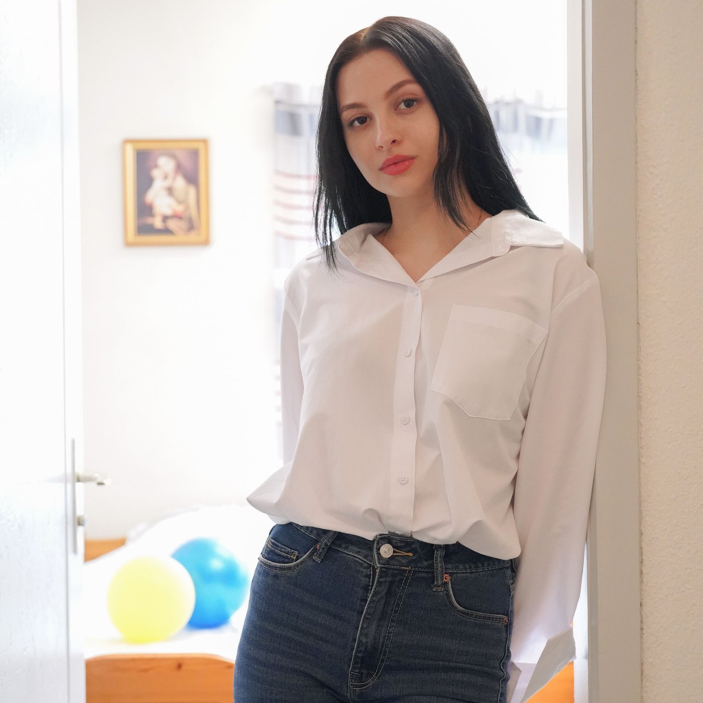 Girl leans against the doorframe in her living room and wears jeans and a white shirt.