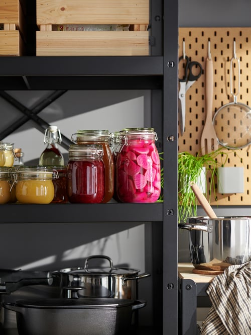 Clear glass KORKEN jars with lids containing pickles and condiments on black BROR shelves holding kitchen utensils and boxes.