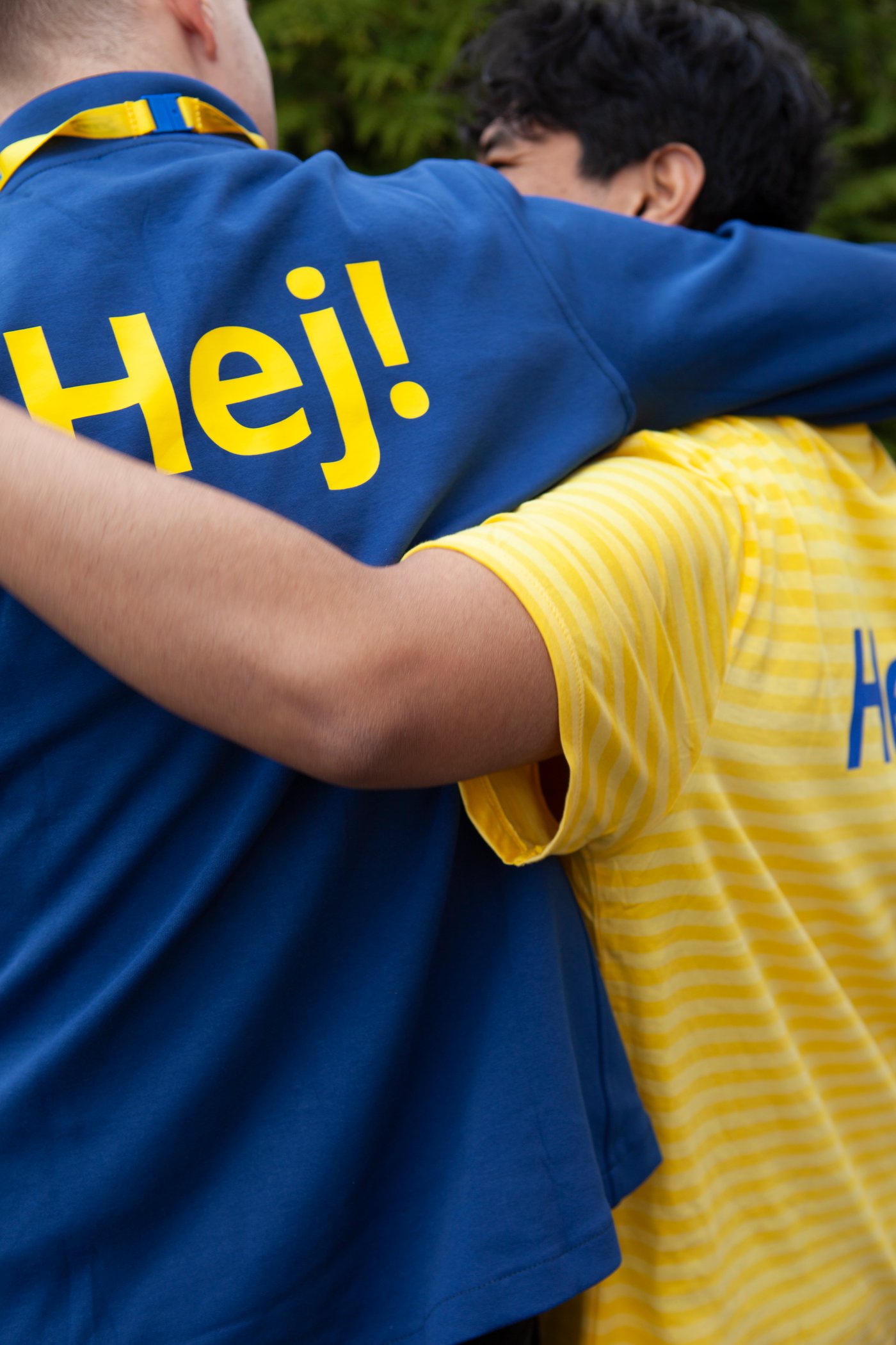 Kaksi työntekijää kävelemässä pitäen toisiaan olkapäistään kiinni. Heidän paidoissaan lukee "Hej!".