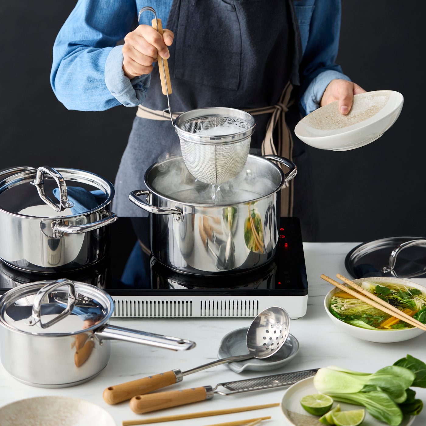 A person cooking on a white TILLREDA portable induction hob with two cooking zones while using a VARDAGEN noodle strainer.