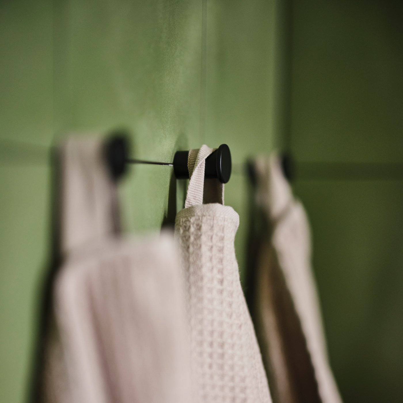 Dark beige towels hanging from three black SKOGSVIKEN hooks against a green tiled wall.