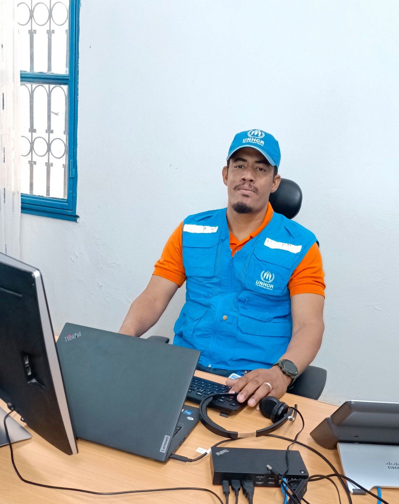 Ilyas wearing his blue UNHCR work uniform sitting by a computer in his office.