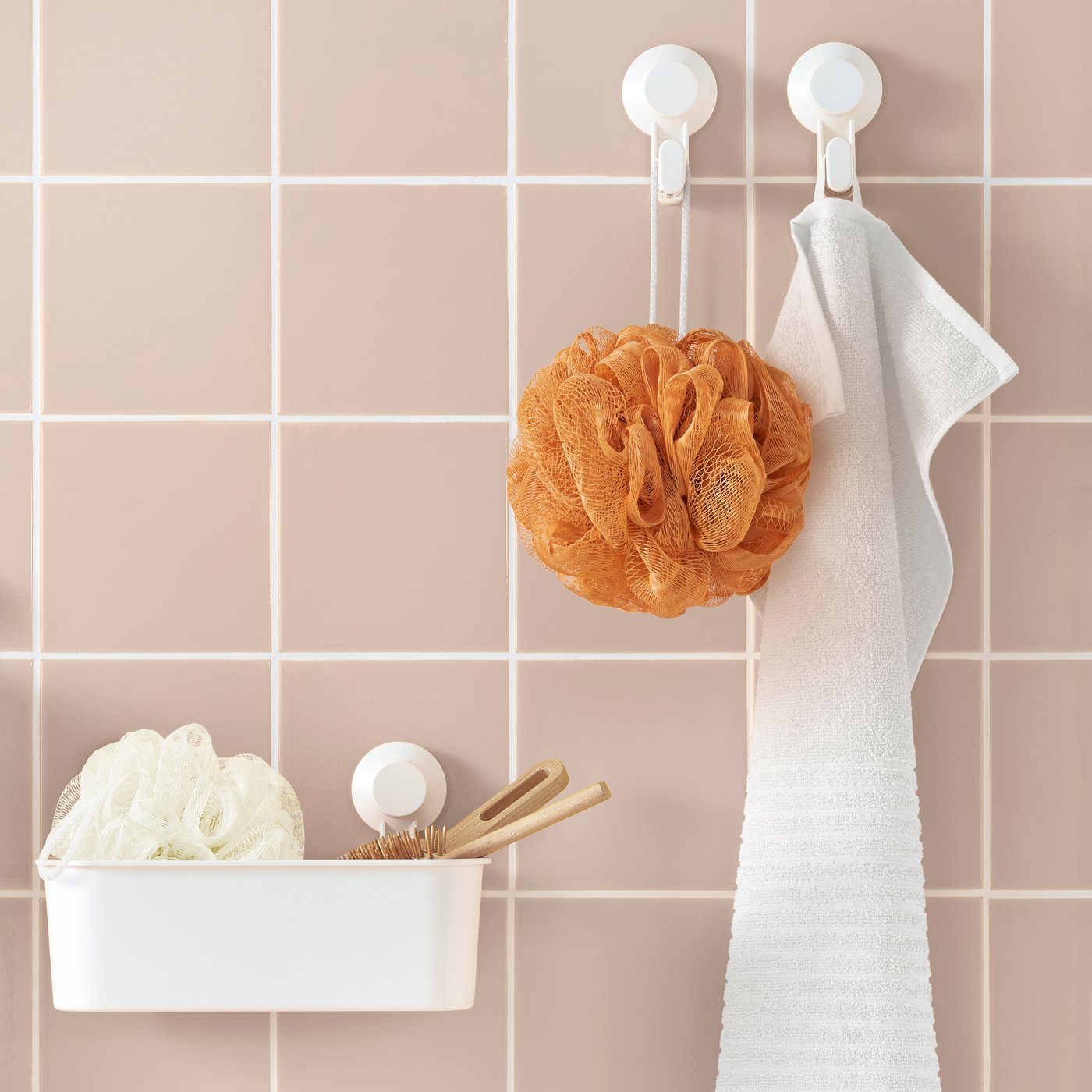 Badezimmerwand mit rosa Fliesen, zwei weißen Haken mit einem orangefarbenen Duschschwamm und einem hängenden Handtuch, daneben ein weißes Regal mit einem weißen Duschschwamm und einer Badbürste aus Holz.