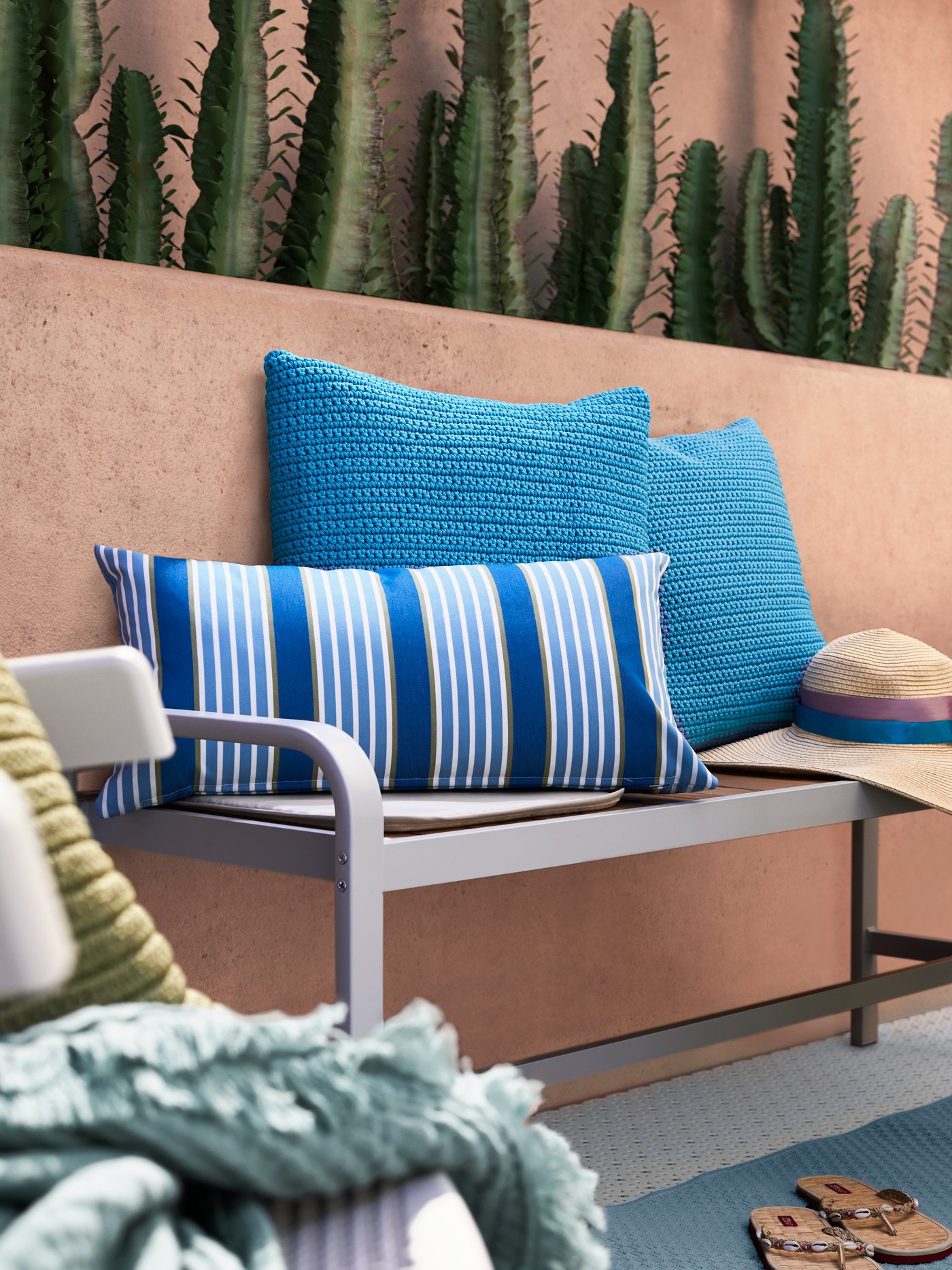 Kusyen corak berjalur-jalur biru, dua kusyen yang dikait berwarna biru dan topi sinar di atas bangku panjang yang bersandar pada dinding terracotta yang mempunyai pokok kaktus.